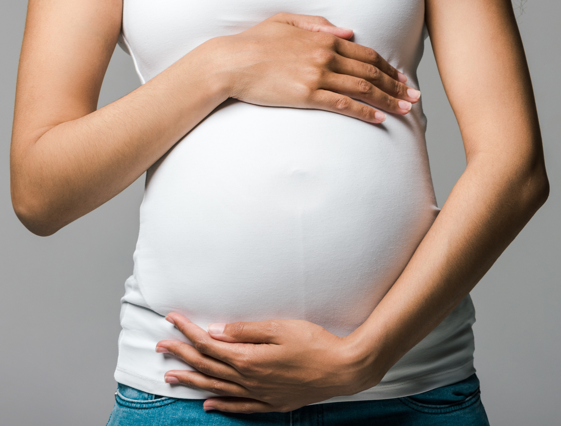 A person showing pregnancy in a white shirt frames in their belly with one arm on top and one on the bottom.