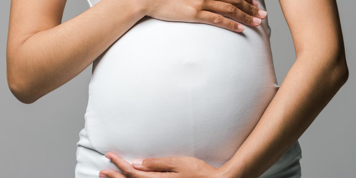 A person showing pregnancy in a white shirt frames in their belly with one arm on top and one on the bottom.